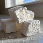 Close-up of four fluffy homemade marshmallows dusted heavily with powdered sugar on a light surface.