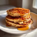 A tall stack of fluffy oatmeal pancakes generously drizzled with golden syrup on a white plate.