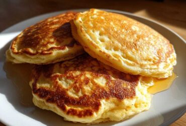 A stack of three golden brown, fluffy oatmeal pancakes drizzled with syrup on a light gray plate.
