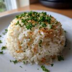A molded serving of fluffy white rice pilaf garnished generously with chopped fresh parsley and crispy fried onions or garlic.