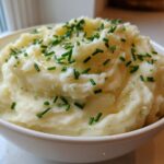 A close-up of a white bowl filled with fluffy garlic mashed potatoes, topped with melted butter and fresh chives.