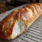 A golden brown, scored loaf of homemade french bread with a bite taken out, resting on a wire cooling rack.
