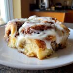 A close-up shot of a fluffy, freshly baked cinnamon roll generously covered in white cream cheese icing.