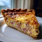 A golden-brown slice of Ham and Cheese Quiche with visible chunks of ham in the creamy egg filling.
