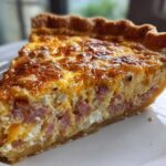 A close-up view of a perfectly baked slice of ham and cheese quiche showing the flaky crust and rich egg filling.