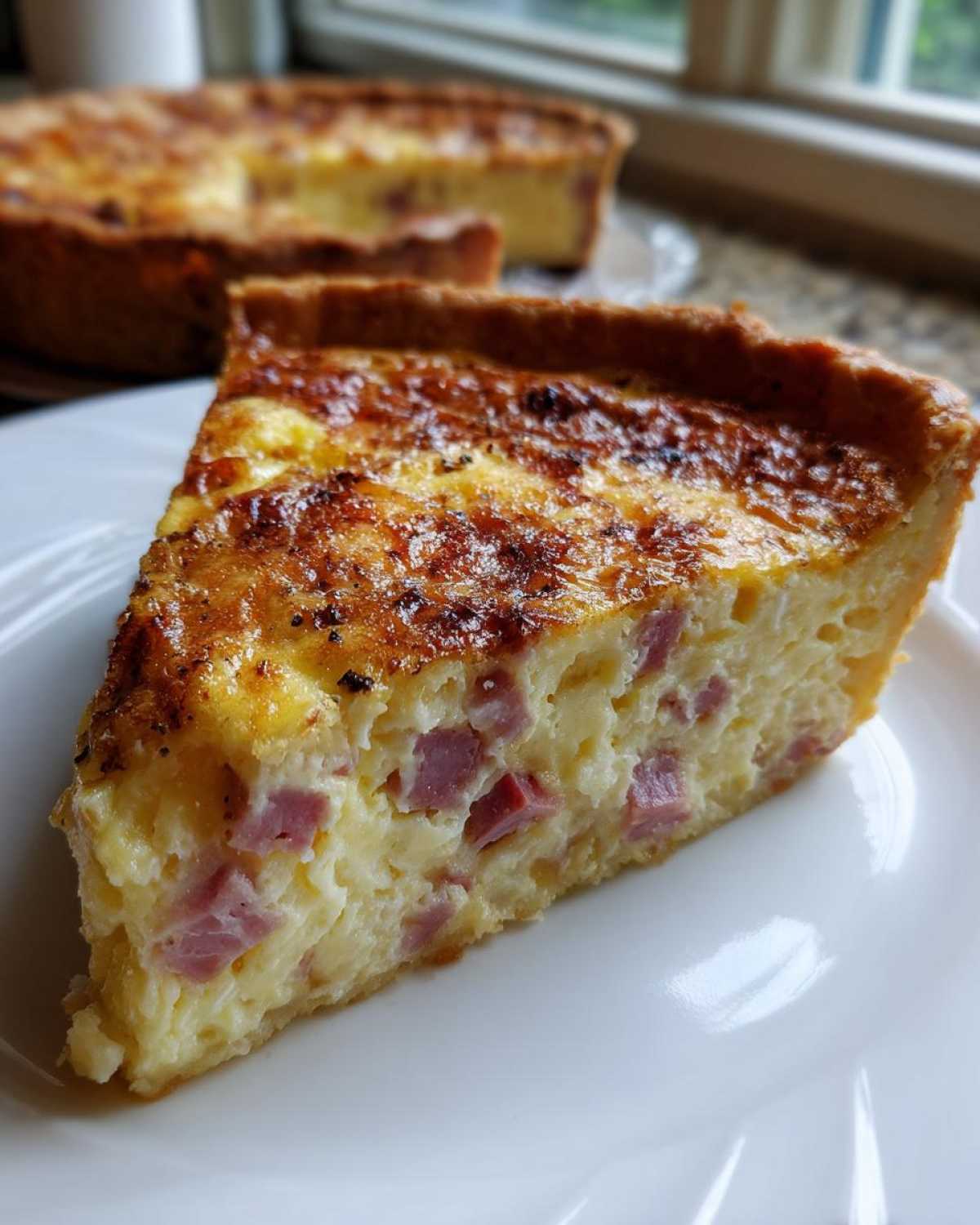 A close-up slice of ham and cheese quiche on a white plate, showing chunks of ham in the creamy filling.