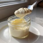 A spoonful of thick, creamy homemade mayonnaise being lifted from a small glass jar.