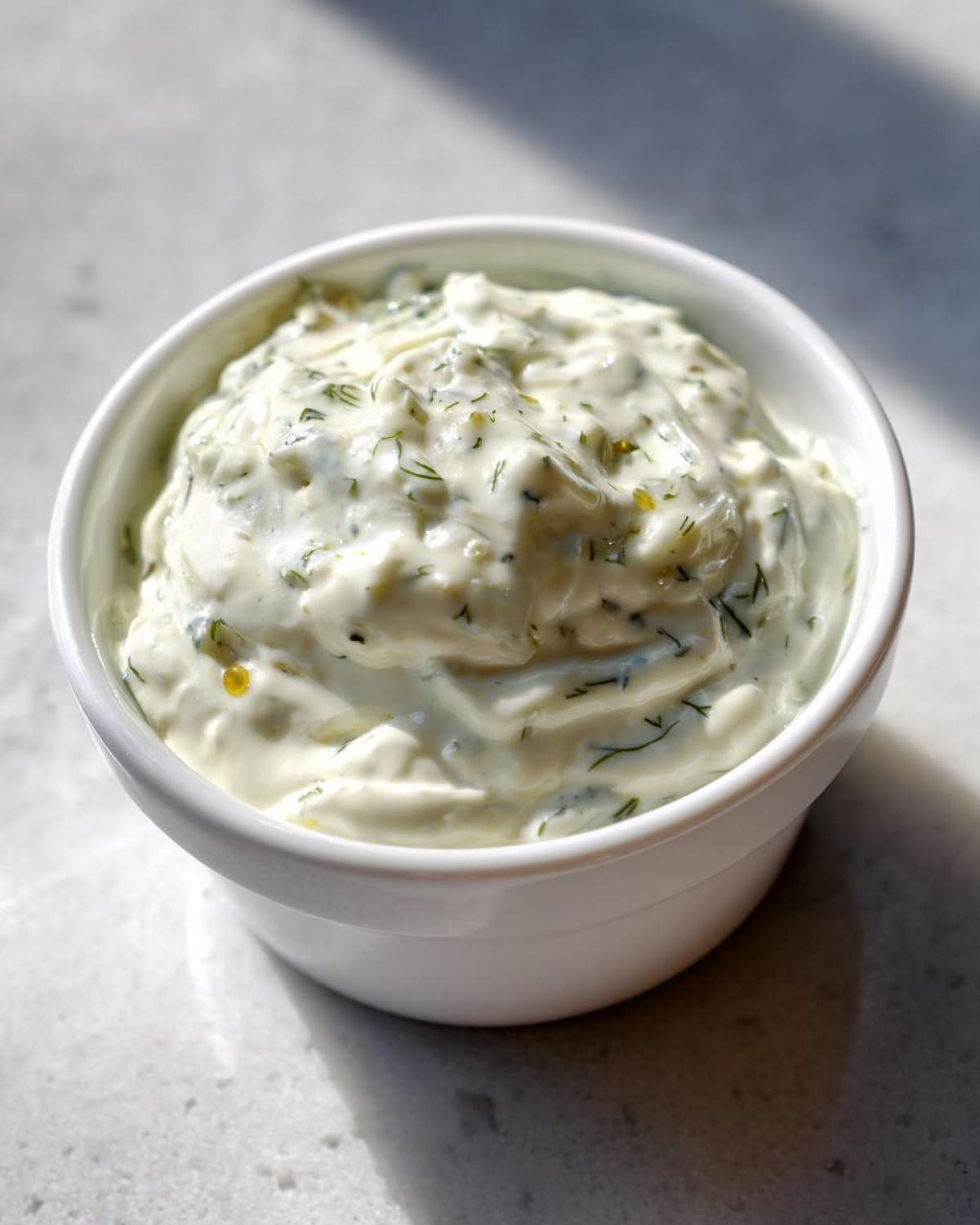 Close-up of creamy, chunky homemade tartar sauce recipe with visible herbs in a small white bowl.