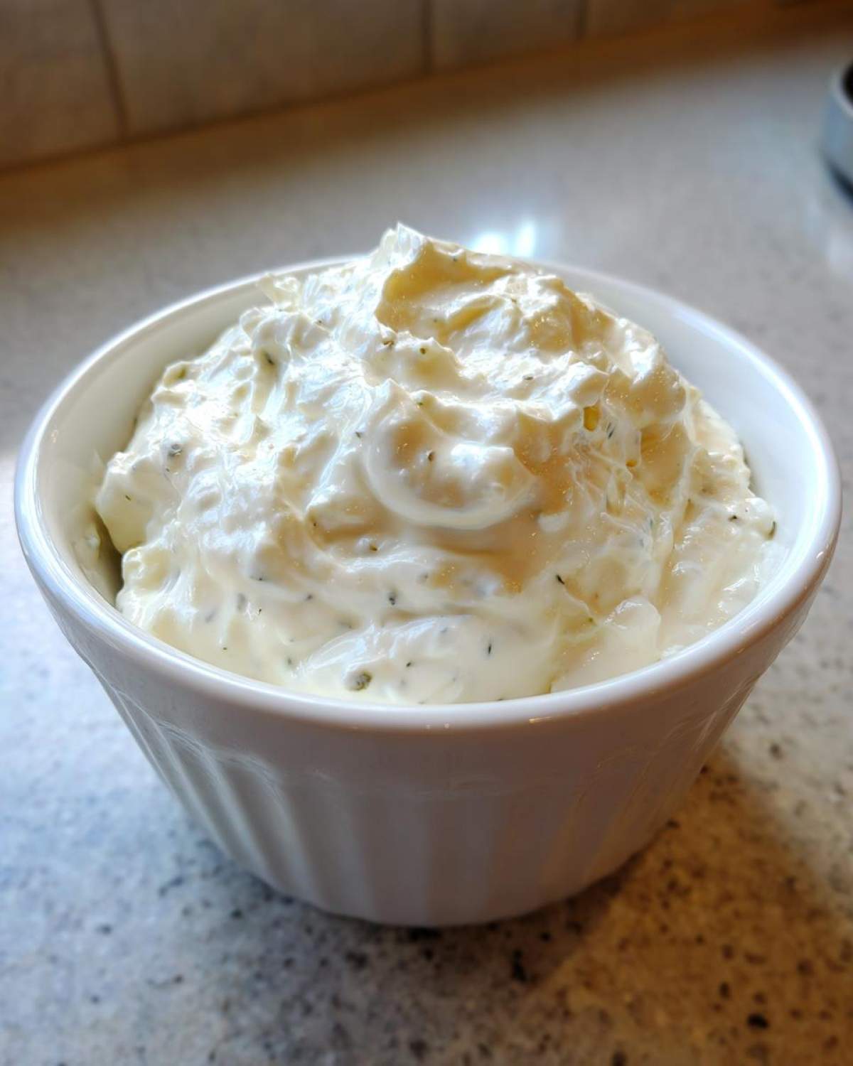 A close-up of creamy, homemade tartar sauce recipe mixture with visible herbs in a small white bowl.