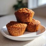 Three golden brown honey bran muffins stacked on a white plate, catching the sunlight.