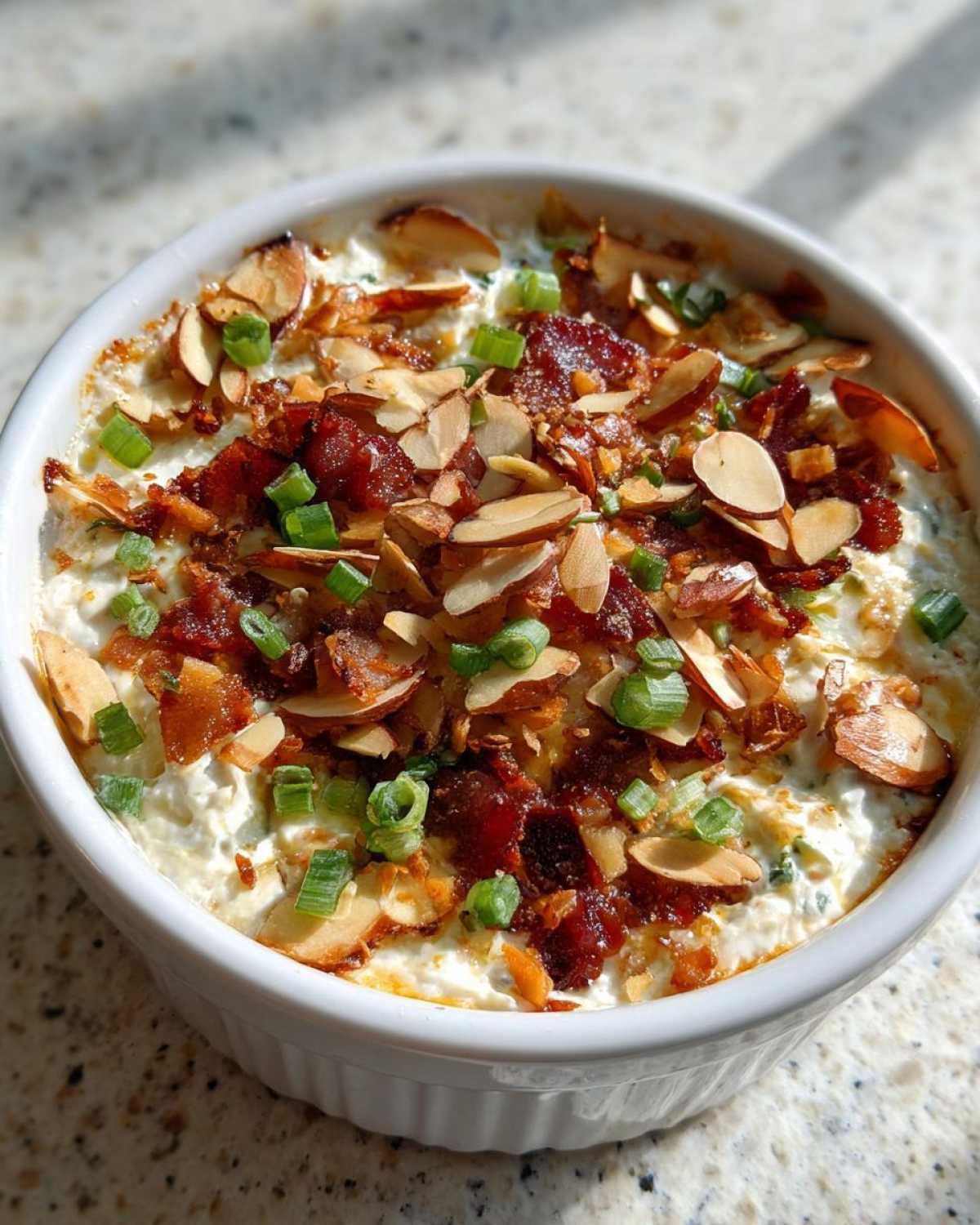 Close-up of hot Neiman Marcus dip in a white ramekin, topped with crumbled bacon, sliced almonds, and green onions.