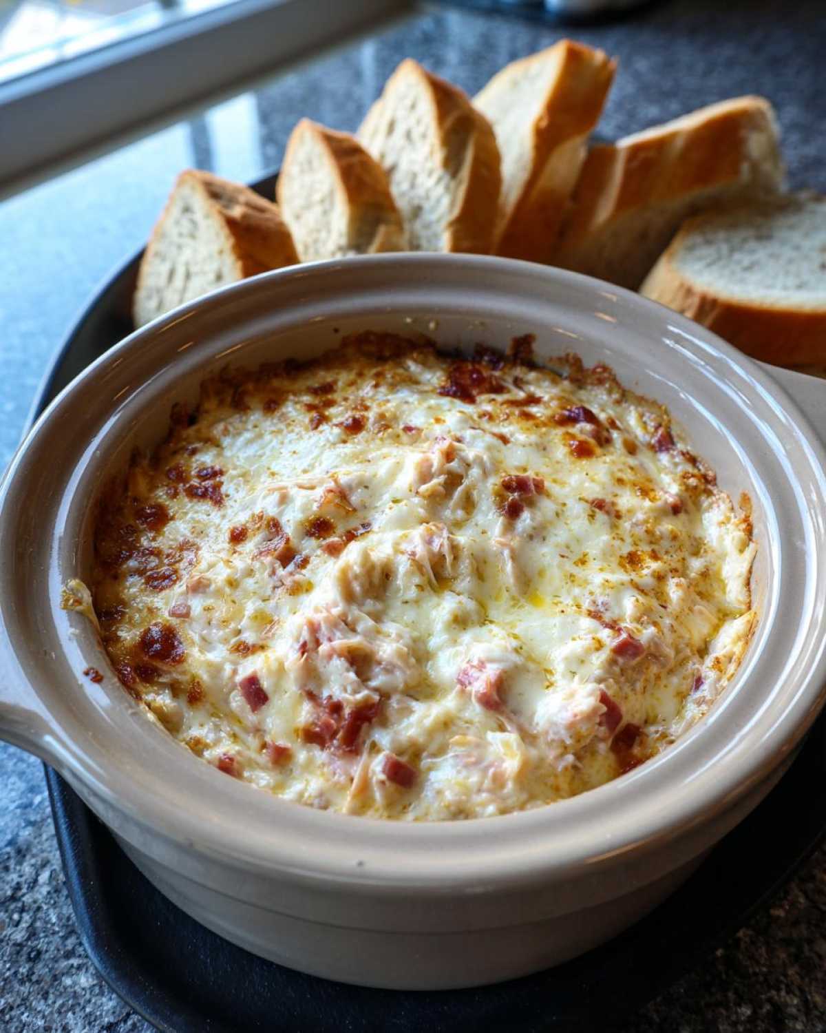 A close-up of a baked, cheesy reuben dip served hot in a ceramic dish with slices of rye bread arranged behind it.