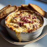 Close-up of a bubbling, hot reuben dip topped with corned beef, served in a white crock with toasted rye bread slices.