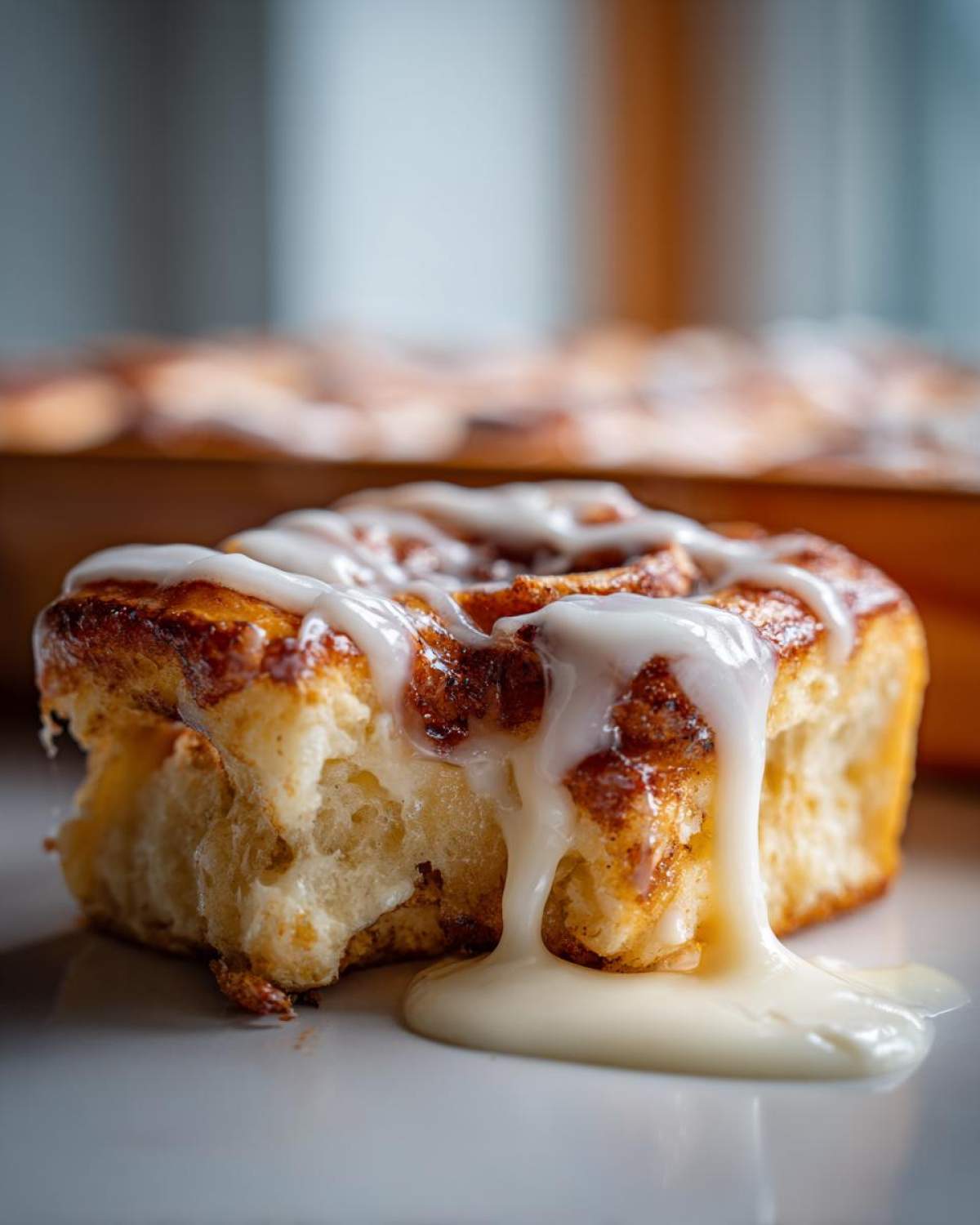 A close-up shot of a soft, baked cinnamon roll with thick white icing dripping down the side onto a white surface.