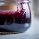 Close-up of dark purple elderberry syrup recipe liquid with vibrant red foam clinging to the inside of a small glass jar.