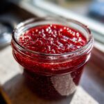 Close-up of a glass jar filled with glistening, deep red homemade raspberry jam, catching the sunlight.