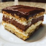 A square slice of layered eclair cake featuring graham crackers, vanilla, and chocolate pudding layers, topped with chocolate frosting.