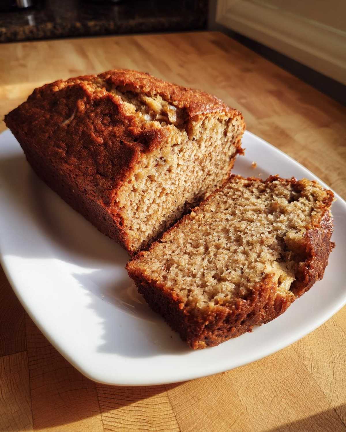 A moist loaf cake, likely an applesauce cake, sliced on a white rectangular plate bathed in sunlight.
