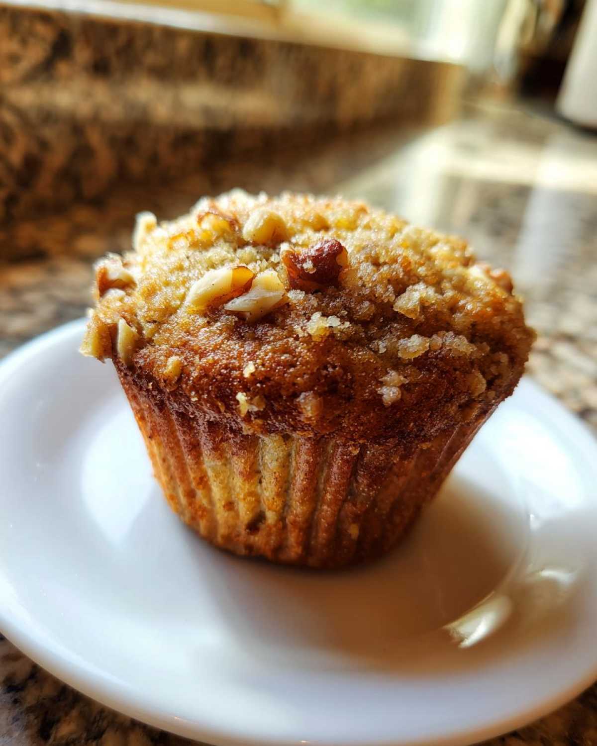 A single, perfectly baked banana nut muffin with a crunchy streusel topping sits on a small white plate.