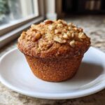 A single, moist banana nut muffin topped with crunchy walnuts, sitting on a small white plate.