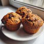 Three freshly baked, moist bran muffins studded with raisins sitting on a white plate near a window.