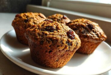 Four moist bran muffins topped with raisins sitting on a white plate, highlighted by natural window light.