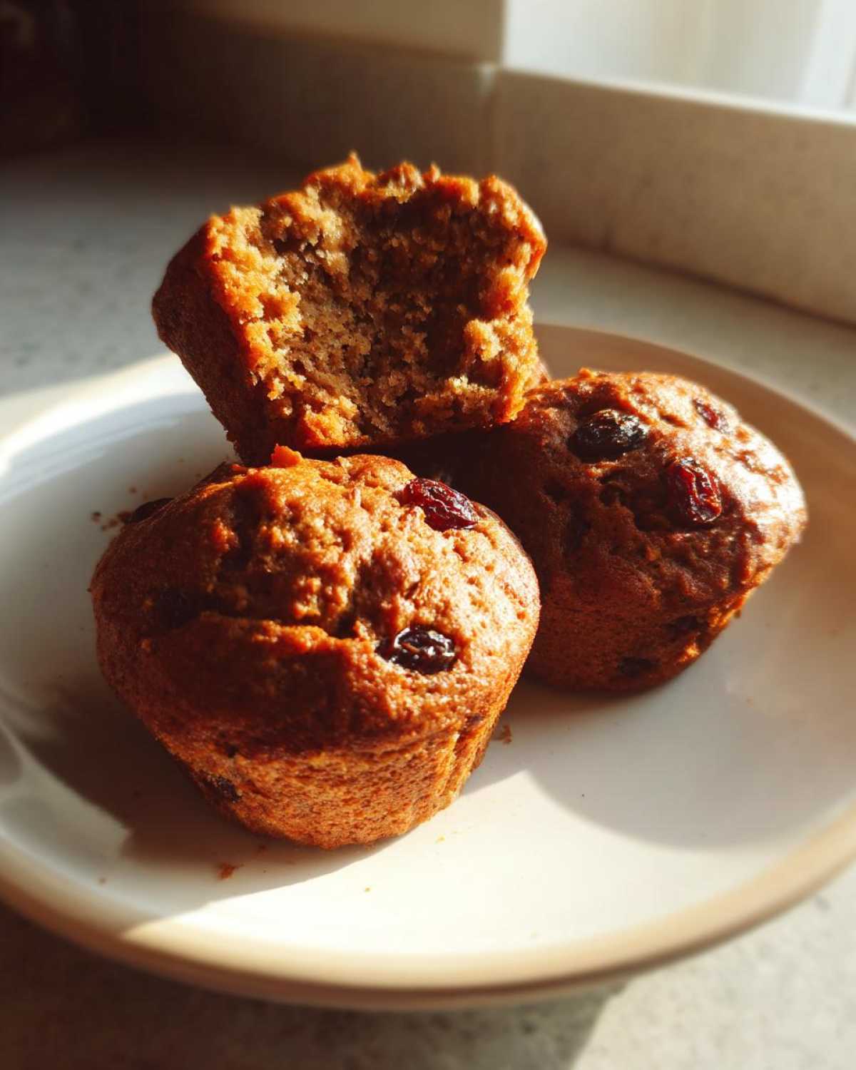 Three moist bran muffins, one broken open to show the texture, sitting on a light plate in bright sunlight.
