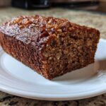 A close-up of a thick slice of rich, dark date nut bread with a glossy top, served on a white plate.