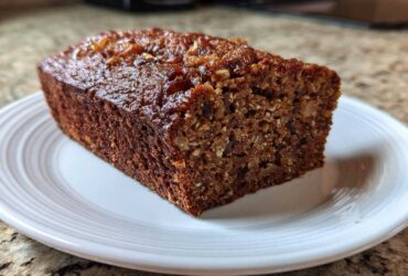 A close-up of a thick slice of rich, dark date nut bread with a glossy top, served on a white plate.