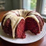 A moist red velvet bundt cake with a thick, creamy glaze drizzled over the top, with one slice cut out.