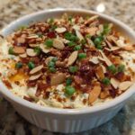 Close-up of a white bowl filled with creamy Neiman Marcus Dip, topped generously with bacon bits, sliced almonds, and green onions.