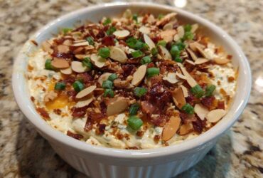 Close-up of a white bowl filled with creamy Neiman Marcus Dip, topped generously with bacon bits, sliced almonds, and green onions.