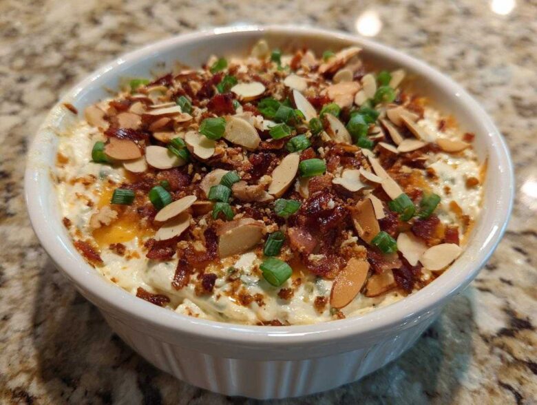 Close-up of a white bowl filled with creamy Neiman Marcus Dip, topped generously with bacon bits, sliced almonds, and green onions.