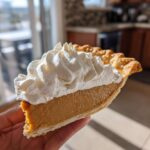 A hand holding a thick slice of creamy butterscotch pie topped generously with whipped cream.