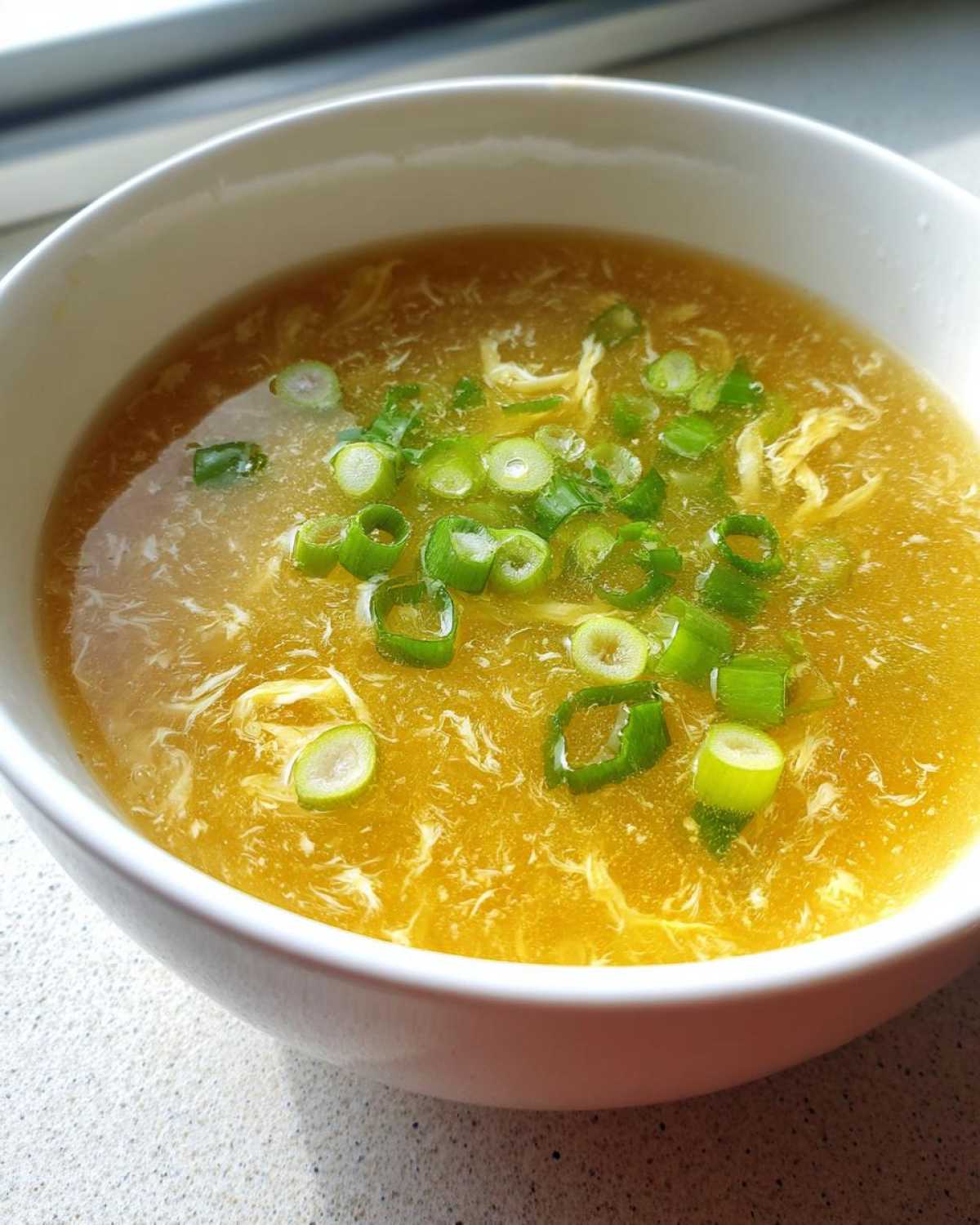 A close-up view of bright yellow egg drop soup in a white bowl, garnished generously with sliced green onions.