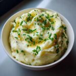 A close-up of a white bowl filled with silky garlic mashed potatoes, topped with melted butter and fresh chopped chives.