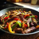 Close-up of sizzling steak fajitas with colorful bell peppers and onions in a dark skillet.