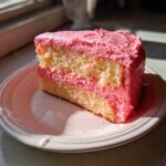 Close-up of a moist slice of strawberry cake with bright pink frosting and filling, sitting on a light pink plate.