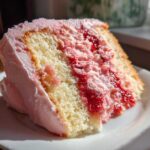 Close-up of a moist slice of strawberry cake with layers of vanilla sponge, pink frosting, and bright red strawberry filling.