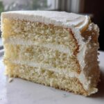 Close-up of a moist, three-layer slice from a vanilla cake recipe, generously frosted with white buttercream.