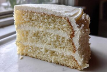Close-up of a moist, three-layer slice from a vanilla cake recipe, generously frosted with white buttercream.
