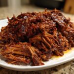 A close-up of a generous mound of saucy, shredded slow cooker pulled pork piled high on a white serving dish.