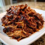 A close-up of tender, shredded slow cooker pulled pork coated in a rich, glossy barbecue sauce on a white plate.
