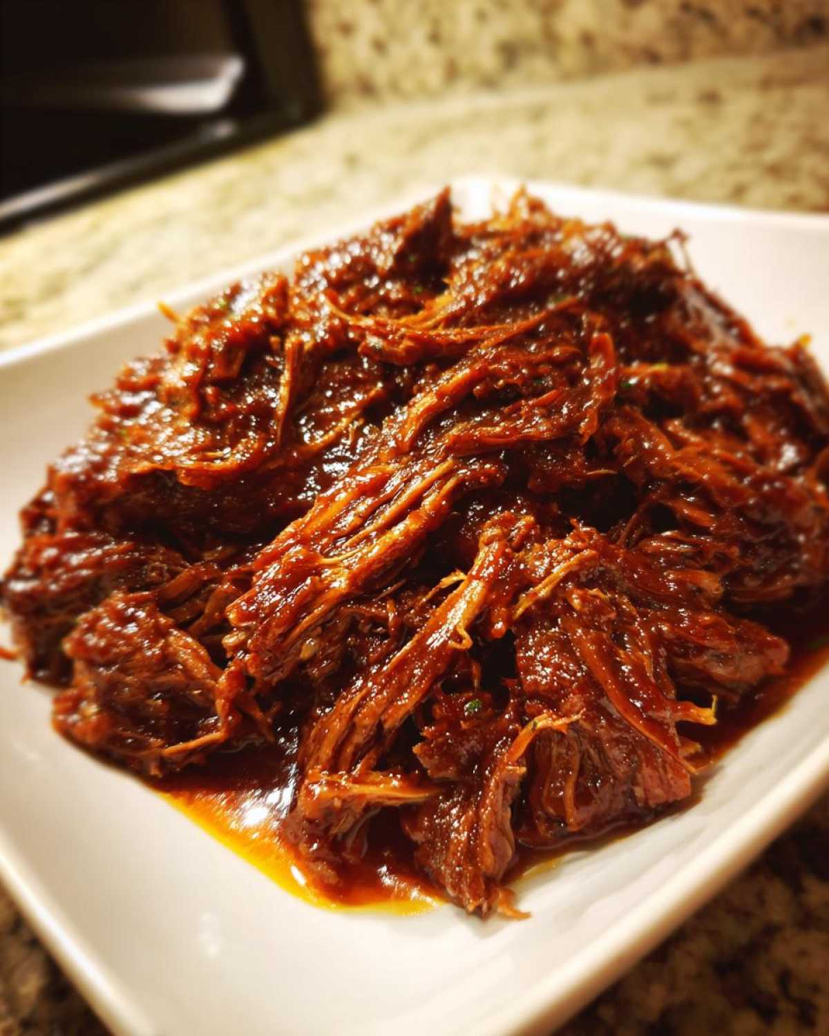 Close-up of richly sauced, shredded slow cooker pulled pork piled high on a white square plate.