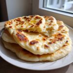 A stack of freshly made, soft and chewy homemade naan bread, lightly brushed with butter, resting on a white plate.
