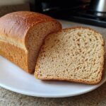 A freshly baked loaf of soft whole wheat bread with one slice resting against it on a white plate.