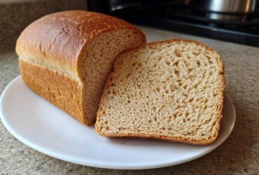A freshly baked loaf of soft whole wheat bread with one slice resting against it on a white plate.