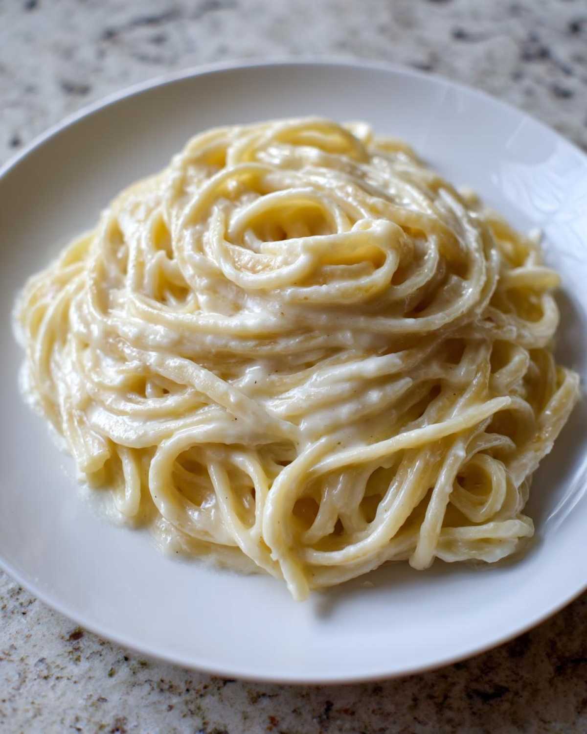 A close-up of spaghetti noodles generously coated in rich, creamy homemade alfredo sauce on a white plate.