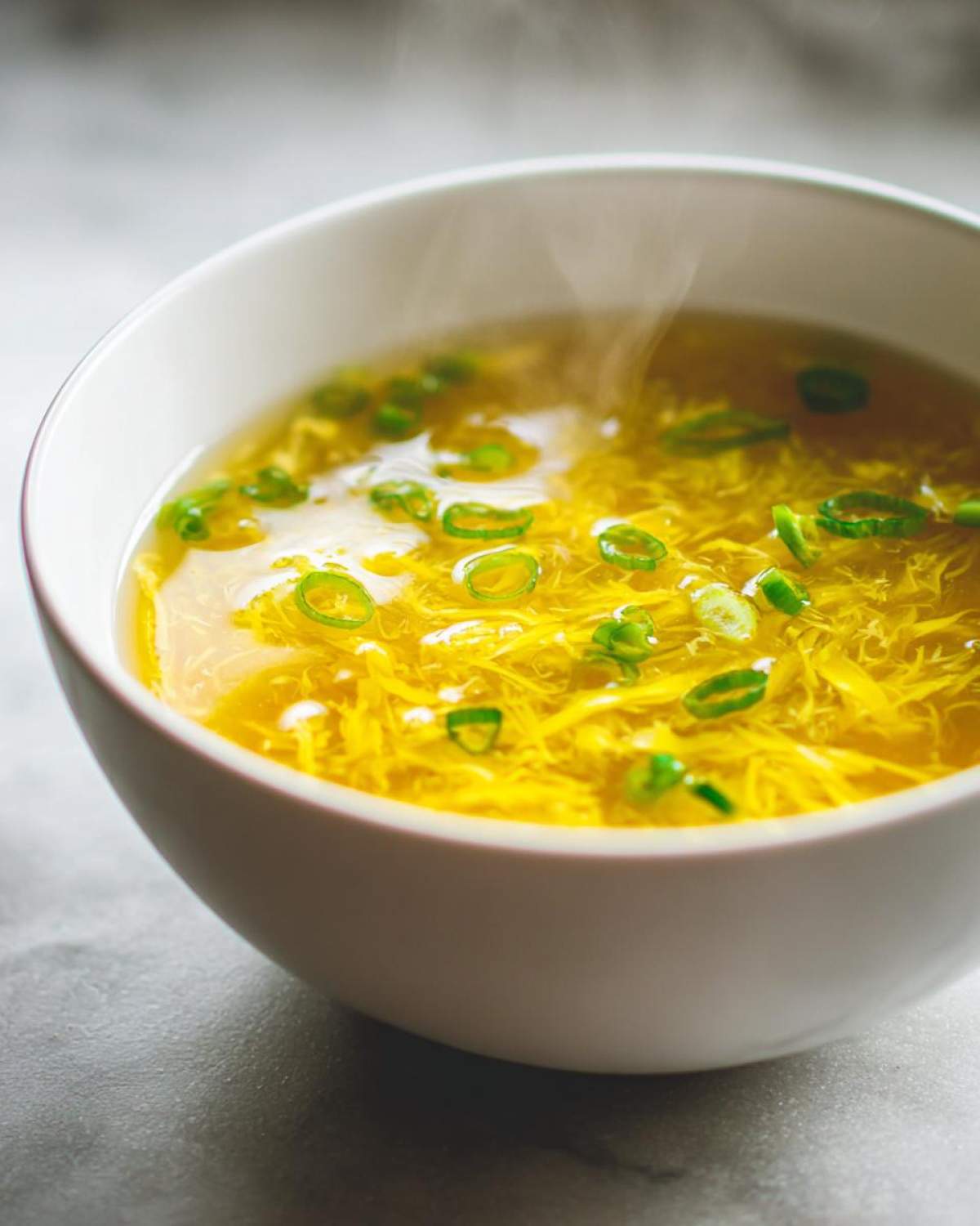 Close-up of a steaming white bowl filled with bright yellow egg drop soup garnished with sliced green onions.