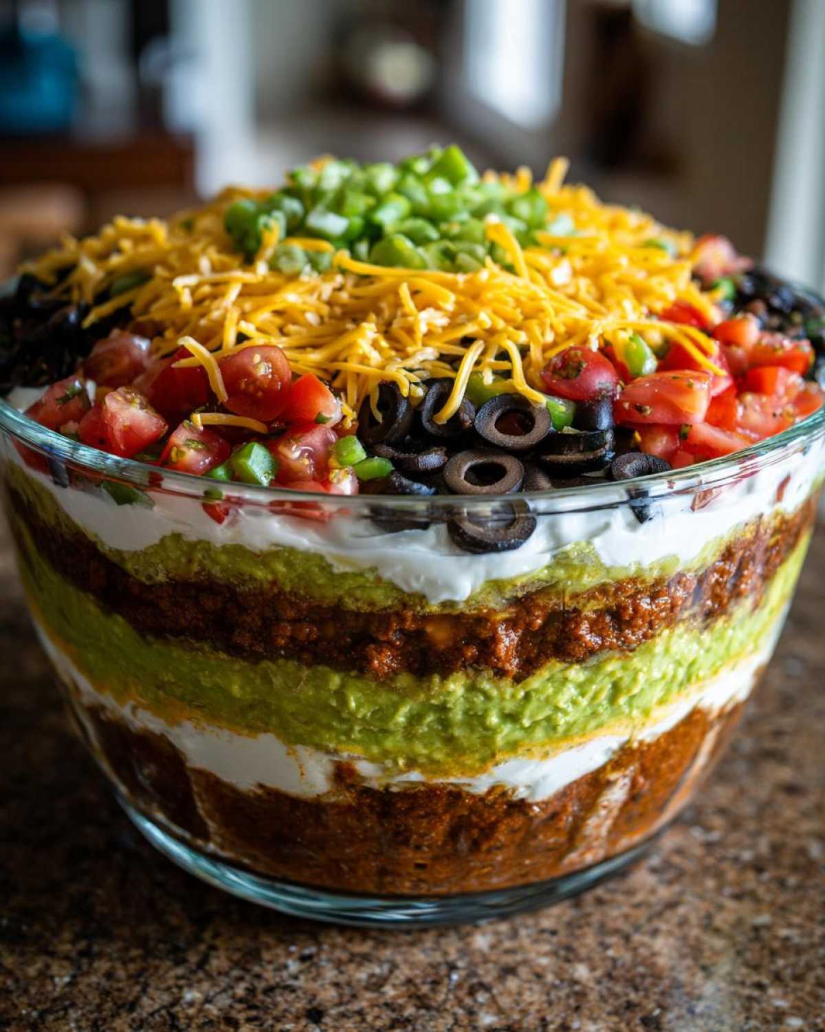 Close-up of a towering Seven layer dip in a clear glass bowl, topped with cheese, olives, tomatoes, and green onions.